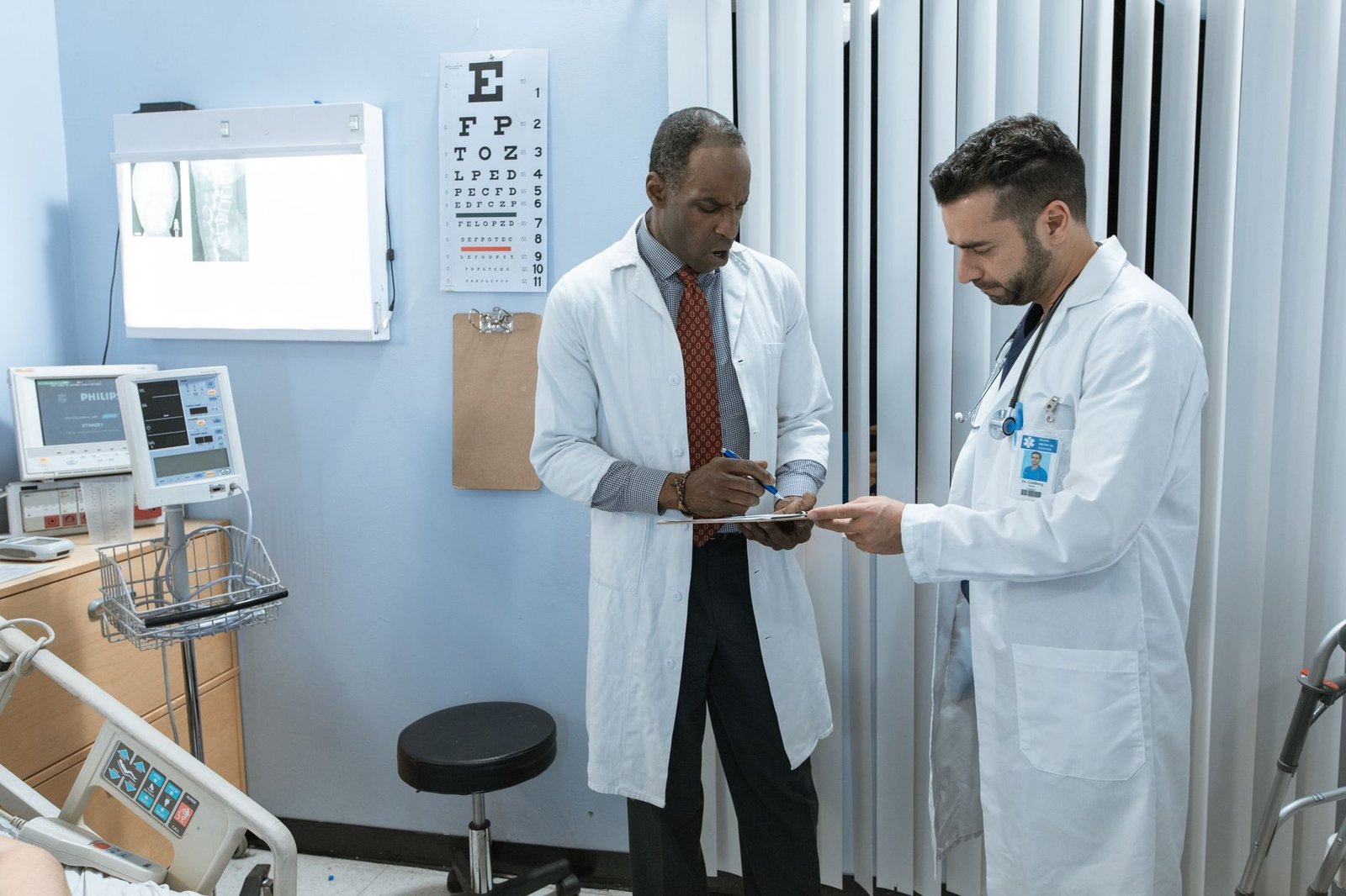 Two clinicians conferring over a patient chart in an exam room, a cervical spine radiograph lit on the viewer behind them.