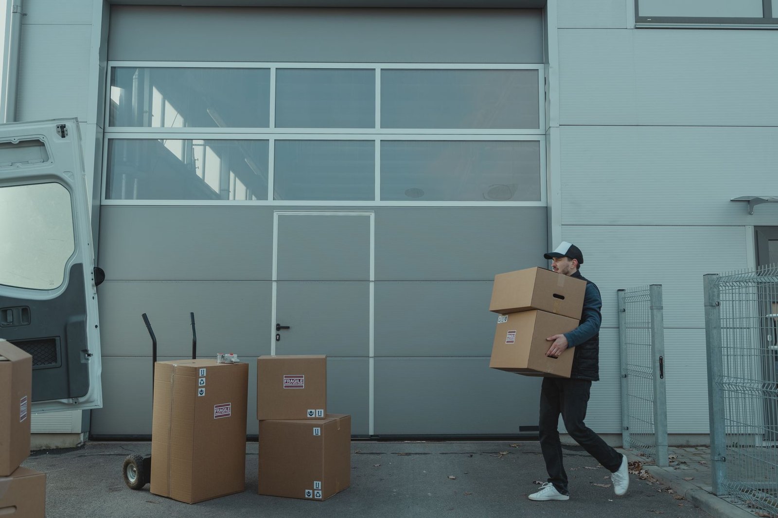 A delivery worker carrying two stacked boxes away from the body outside a loading dock, a load pattern that strains the neck and shoulder.