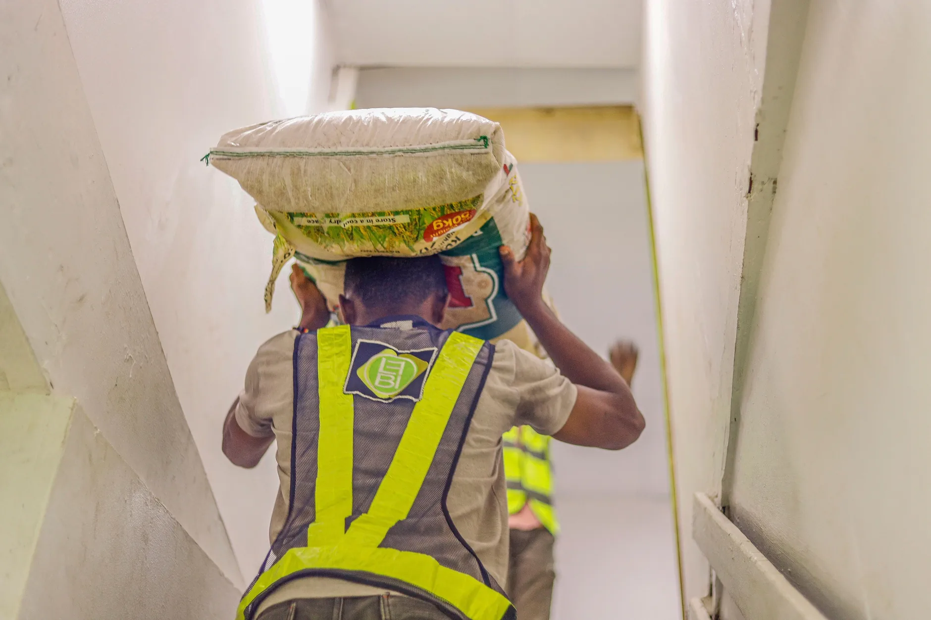 A worker in a safety vest carries a heavy sack on his head up a stairwell, a load that strains the neck