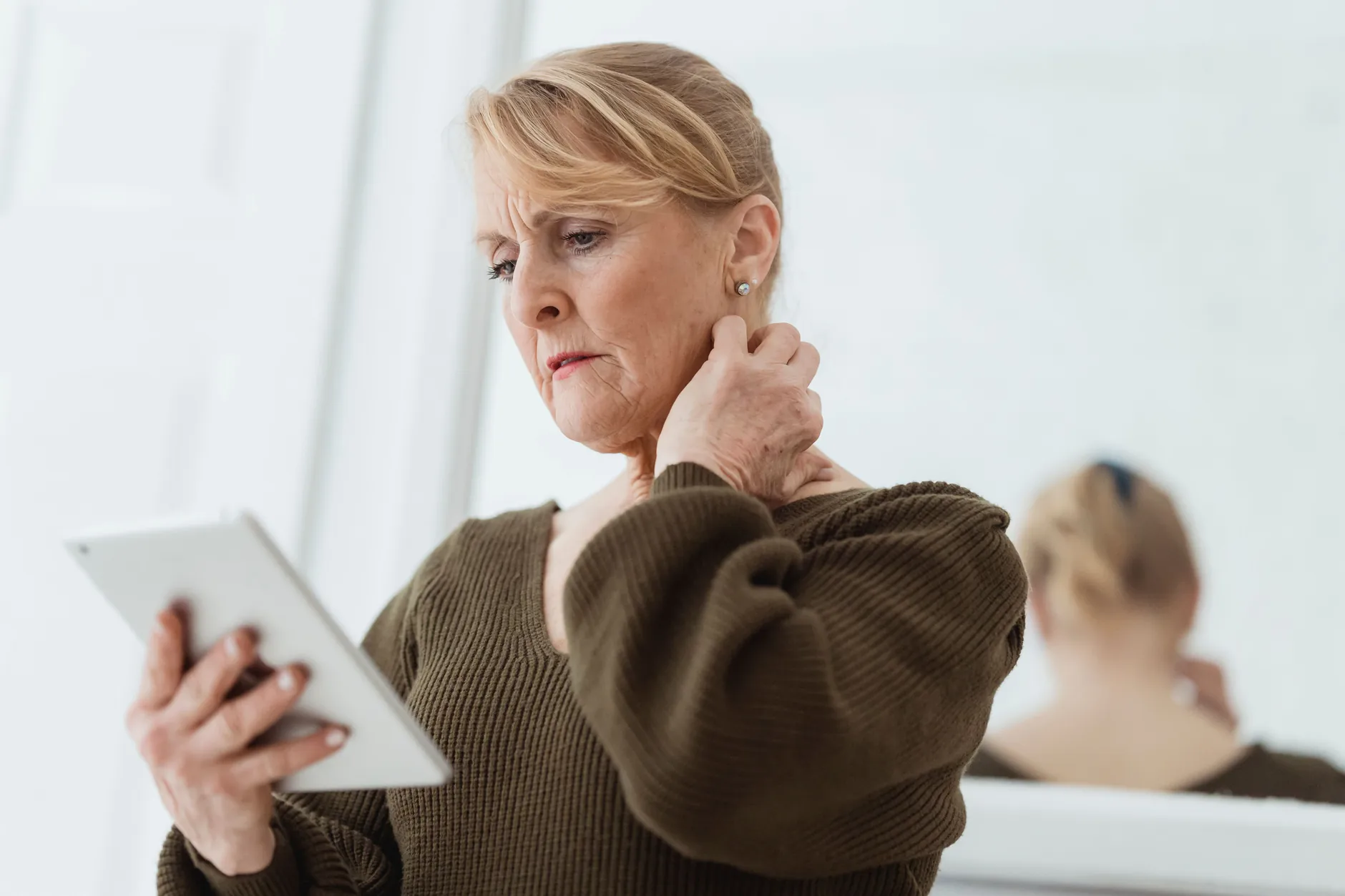 A woman rubs the side of her stiff neck while holding a tablet at home